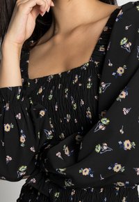 Robe noire ornée de motifs floraux en rose et jaune, corsage smocké, et manches bouffantes avec des poignets élastiques. Tissu doux et léger.