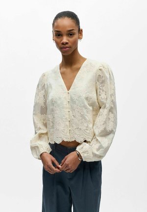 Frau trägt eine cremefarbene Bluse aus floraler Spitze mit aufgeblähten langen Ärmeln und dunkle, hoch taillierte Hose, steht vor weißem Hintergrund.