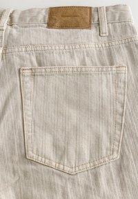 Bežové denimové nohavice s vrúbkovaným vzorom, s jednou zadnou vreckom a hnedou koženou nášivkou na páse, na ktorej je napísané "DENIM SUPPLY."