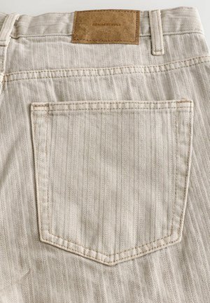 Pantaloni din denim bej, cu un tipar în zigzag, având un buzunar pe spate și o etichetă din piele maro la talie care citește "DENIM SUPPLY."