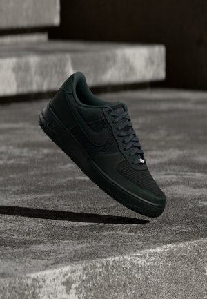 Must Nike Air Force 1 tossud võrgust ja nahast detailidega, hõljuvad tekstureeritud betoonastmete kohal väheses valgustuses.