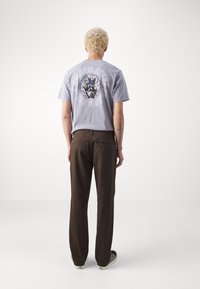 Hellblaues Baumwoll-T-Shirt mit einem bunten Flammenmotiv auf dem Rücken, kombiniert mit braunen geraden Hosen und schwarzen Schuhen.