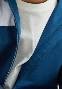 Modrá bunda na zip s hladkou texturou přes bílý tričko. Bunda má kontrastní design a viditelné švy.