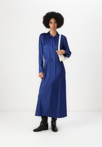 Abito lungo in satin blu profondo con bottoni frontali, vita arricciata e maniche lunghe. Abbinato con stivali neri fino al ginocchio e una borsa leggera.