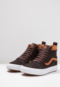 Braune High-Top-Sneaker aus Wildleder mit hellbraunen Akzenten, braunen Schnürsenkeln, weißen Gummisohlen und einer Zuglasche hinten, präsentiert auf einer weißen Oberfläche.