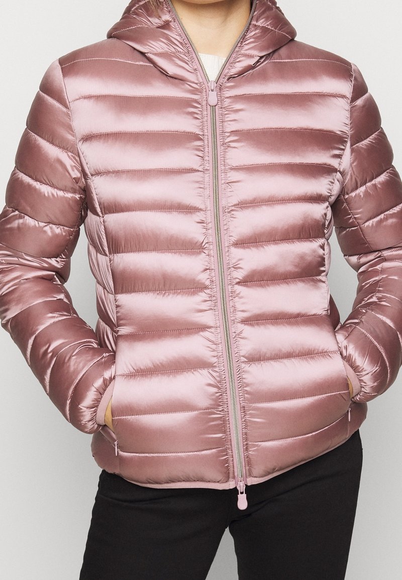 Personne portant une doudoune brillante mauve clair avec un matelassage horizontal, une fermeture éclair à l'avant, une capuche et les mains dans les poches latérales.
