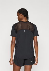 Μαύρο αθλητικό μπλουζάκι με διαφανές πλέγμα στο πίσω μέρος και στρογγυλεμένο τελείωμα, συνδυασμένο με μαύρο σορτς. Απλός σχεδιασμός με ελάχιστες λεπτομέρειες.