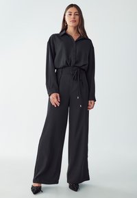 Black, loose-fitting long-sleeve button-up shirt paired with high-waisted wide-leg pants, featuring a tie waist and smooth texture.