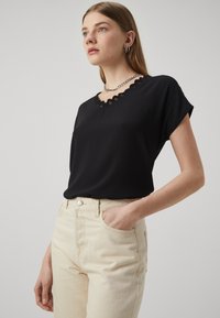 Svart kortermet topp med bølgekant V-hals, laget av myk tekstil. Kombinert med lysebeige høyt liv bukser for kontrast.