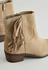 Stivale alla caviglia in suede beige con zip laterale e dettagli in frange. Presenta un tacco basso in legno marrone e cuciture a contrasto.