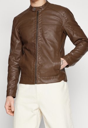 Homme portant une veste en cuir marron avec fermeture éclair et col pressionné, associée à un pantalon blanc, debout avec une main dans la poche.