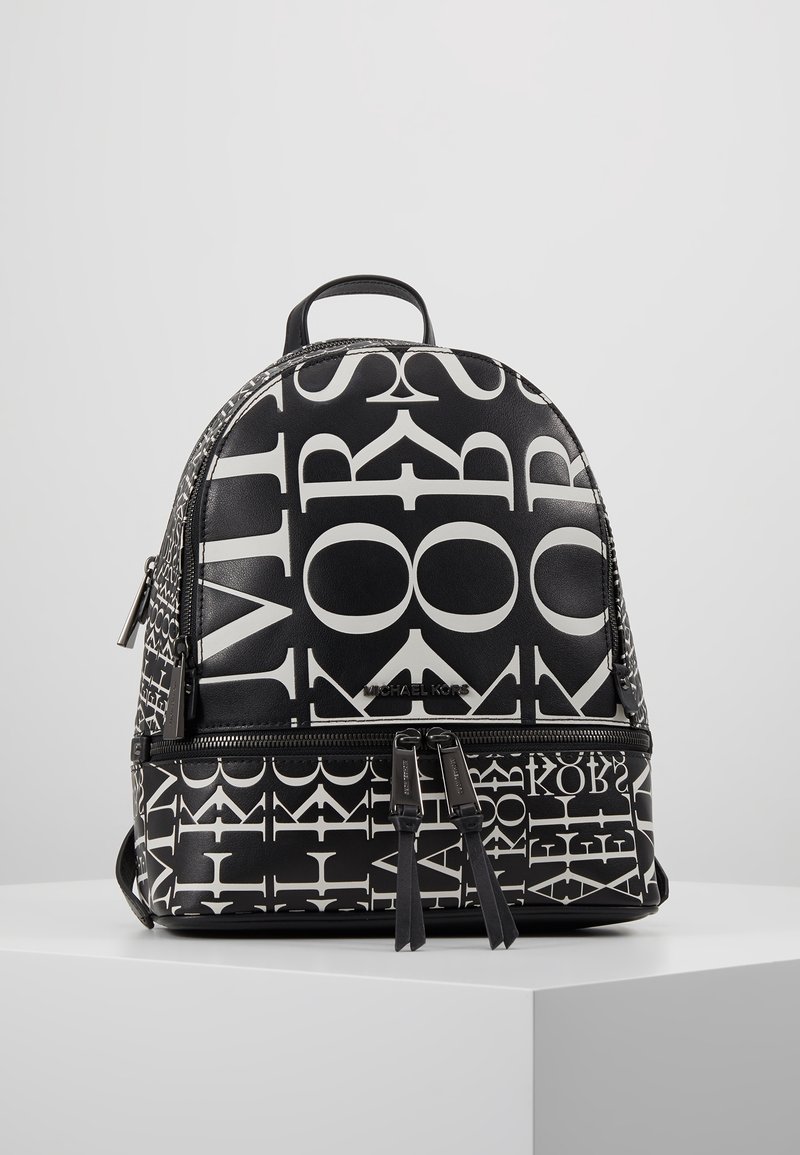 Sac à dos Michael Kors noir et blanc avec un imprimé audacieux de lettres mélangées et deux compartiments zippés, présenté sur un socle blanc.