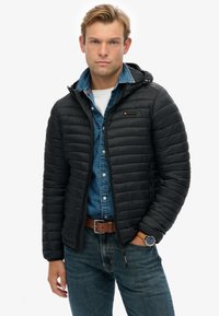 Sort dunjakke med vandret quiltet design, der har en hætte, frontlynlås og logo-detalje. Båret over en denimskjorte og blå jeans.