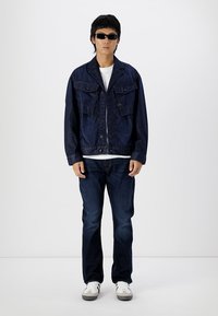 Jachetă din denim de culoare albastru închis, cu două buzunare la piept și închidere cu nasturi, asociată cu blugi închisi. Accesoriile includ ochelari de soare subțiri și adidași.