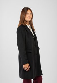 Giovane donna con capelli castani lisci indossa un cappotto nero su una camicia bianca e pantaloni bordeaux, in piedi contro uno sfondo grigio chiaro uniforme.