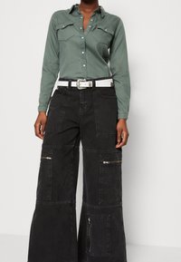 Vihreä pitkähihaisten nappipaitojen, joissa on kaksi rintataskua, yhdistetty mustiin leveälahkeisiin cargo-housuihin, joissa on useita vetoketjutaskuja. Valkea vyö kuuluu mukaan.