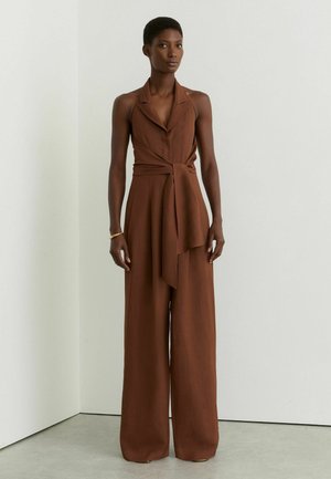 Kobieta stoi, ubrana w bezrękawnikowy brązowy kombinezon z szerokimi nogawkami, kołnierzem i paskiem w talii, na tle białej, gładkiej ściany.