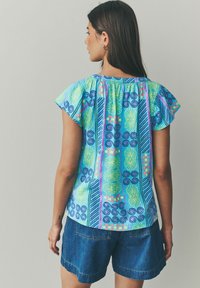 Haut coloré à motifs avec des designs bleu, vert et rose, présentant des manches volantées et un col froncé, associé à un short en denim.