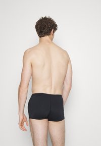 Sorte badeshorts lavet af glat stof, der har en tætsiddende pasform og minimal sømdetaljer, set fra ryggen med en ensfarvet baggrund.
