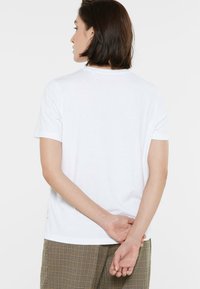 Weißes T-Shirt mit kurzen Ärmeln, rundem Ausschnitt und glatter Textur. Die Rückansicht zeigt ein Etikett am Saum. Kombiniert mit karierten Hosen.