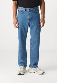 Lösa blå denimjeans med hög midja, kontrasterande sömmar och knappstängning. Bärs med ljusa sneakers.