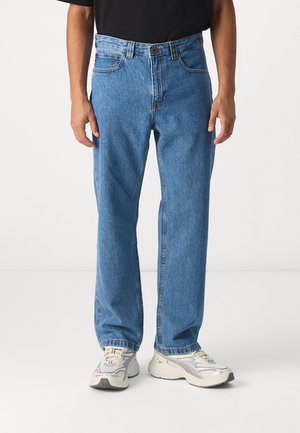 Jean en denim bleu à taille haute, coupe ample, avec des coutures contrastées et un bouton de fermeture. Porté avec des baskets de couleur claire.