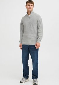 Sudadera de punto gris con cuello con cremallera, combinada con jeans de mezclilla azul oscuro y zapatillas grises. Presenta un tejido texturizado y un corte relajado.