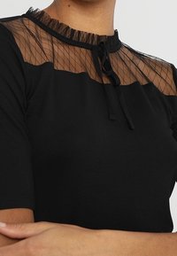 Sort top med en gennemsigtig mesh yoke med gittermønster, en flæsekrave og et sort bånd som accent. Korte ærmer fuldender designet.