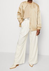 Chaqueta bomber de crema, top de punto a rayas beige, pantalones de pierna ancha de rayas blancas y botas de tobillo puntiagudas en crema. Tejido texturizado con un brillo suave.