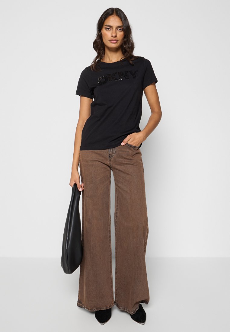 Camiseta de algodón negra con el logo "DKNY" en un diseño texturizado, combinada con unos pantalones vaqueros de pierna ancha marrones y un bolso negro.