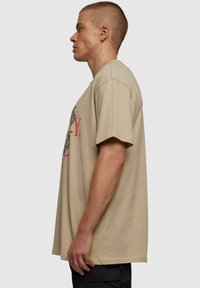 Beige kokvilnas T-krekls ar īsām piedurknēm, brīvs piegriezums, priekšpusē ar grafisku apdruku sarkanā un melnā krāsā, redzamām sānu šuvēm.