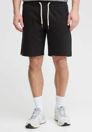 Black sweat shorts with a drawstring waist and side pockets. Cotton fabric, tapered fit, and a small branded label on the lower left side.