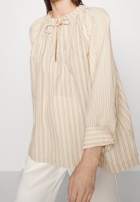 Blouse légère rayée en beige et crème avec un col froncé, une ouverture en goutte d'eau et des manches amples et flottantes. Tissu texturé.