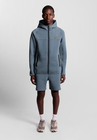 Lichtblauwe hoodie met rits, twee zwarte zijzakken, gecombineerd met lichtblauwe shorts. Gestructureerde stof met een klein geel logo-accent.