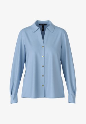 Frau trägt eine hellblaue, geknöpfte Bluse, gemusterte schmale Hose, schwarze klobige Loafers und hält eine kleine schwarze Handtasche am Griff.