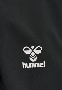 Tessuto nero con un logo bianco raffigurante un'ape stilizzata e la parola "hummel" sottostante, che riflette una texture liscia e atletica.