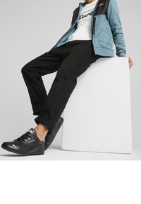 Baskets noires avec accents blancs, pantalon noir retroussé et veste zippée bleu clair par-dessus un t-shirt blanc. Assis sur un bloc blanc.