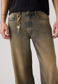 Ξεθωριασμένο μπλε τζιν με ίσια γραμμή στα πόδια. Περιλαμβάνει μπροστινές τσέπες και χρυσό μεταλλικό κλιπ  με κλειδιά προσαρτημένα στη μέση.