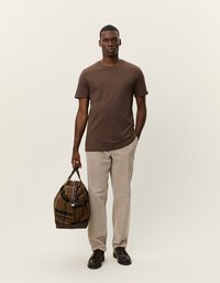 Brun bomulds T-shirt, lige pasform, parret med letvægts beige bukser. Modellen holder en ternet håndtaske og bærer mørke sko.