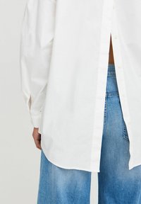 Camicia bianca con bottoni, orlo arrotondato e maniche lunghe, abbinata a pantaloni in denim blu a vita alta con una texture sbiadita.