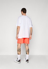 Mann som står med ryggen til, iført en hvit oversized t-skjorte, lyseoransje shorts, hvite sokker, joggesko og synlige tatoveringer på armer og ben.