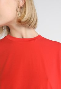 Top rosso a maniche corte con scollatura tonda. Tessuto liscio di un colore uniforme. I capelli del modello sono biondi e acconciati in un caschetto corto. Orecchini a cerchio dorati.
