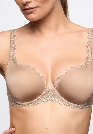 Vrouw draagt een beige beha met kanten afwerking en kleine strikjes op de bandjes en middenvoor, waarbij de bovenste romp en nek zichtbaar zijn.