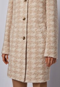 Beige og cremefarvet uldfrakke med tekstur, prydet med skildpaddeskalsknapper og sidelommer, båret over tanfarvede bukser, vist fra midt på låret til hænderne.