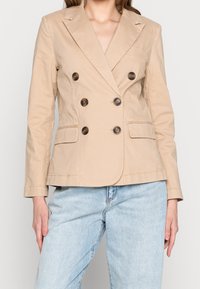 Beżowa marynarka dwurzędowa z klapkami, brązowymi guzikami, dwoma przednimi kieszeniami i dopasowanym krojem, zestawiona z jasnoniebieskimi dżinsami.