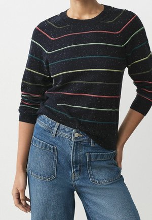 Femme portant un pull bleu marine foncé à rayures horizontales multicolores et un jean denim bleu taille haute avec des poches plaquées à l'avant.