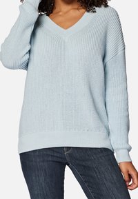 Hellblaues Strickpullover mit V-Ausschnitt, lockerer Passform, gerippten Bündchen und Saum sowie strukturiertem Muster. Getragen mit dunklen Jeans.