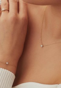 Goldene Halskette mit einem kleinen klaren Edelstein-Anhänger, auf der Haut getragen, und ein goldener Ring mit einem ähnlichen Edelstein an einem Finger. Weißer strukturierter Pullover.