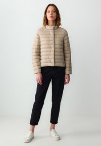 Giubbotto imbottito beige con colletto alto, design a righe orizzontali e chiusura frontale con bottoni a pressione. Indossato con pantaloni neri e sneakers bianche.