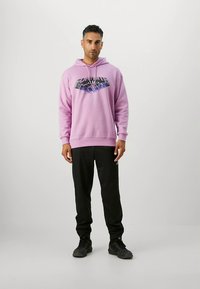 Camisola com capuz lilás claro com estampa gráfica no peito, combinada com calças de fato de treino pretas e sapatilhas pretas. Tecido suave, corte folgado.